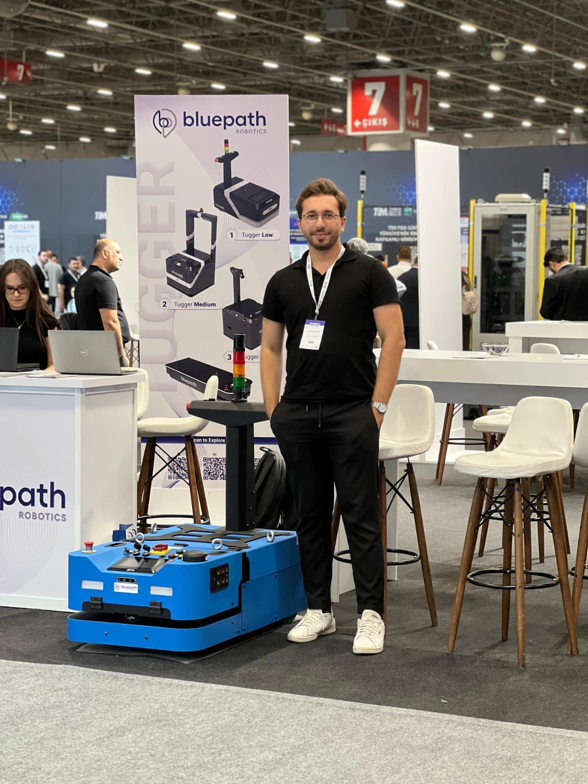 Tolgahan Gergin, bir fuarda Bluepath Robotics standında mavi tugger AMR'ın yanında