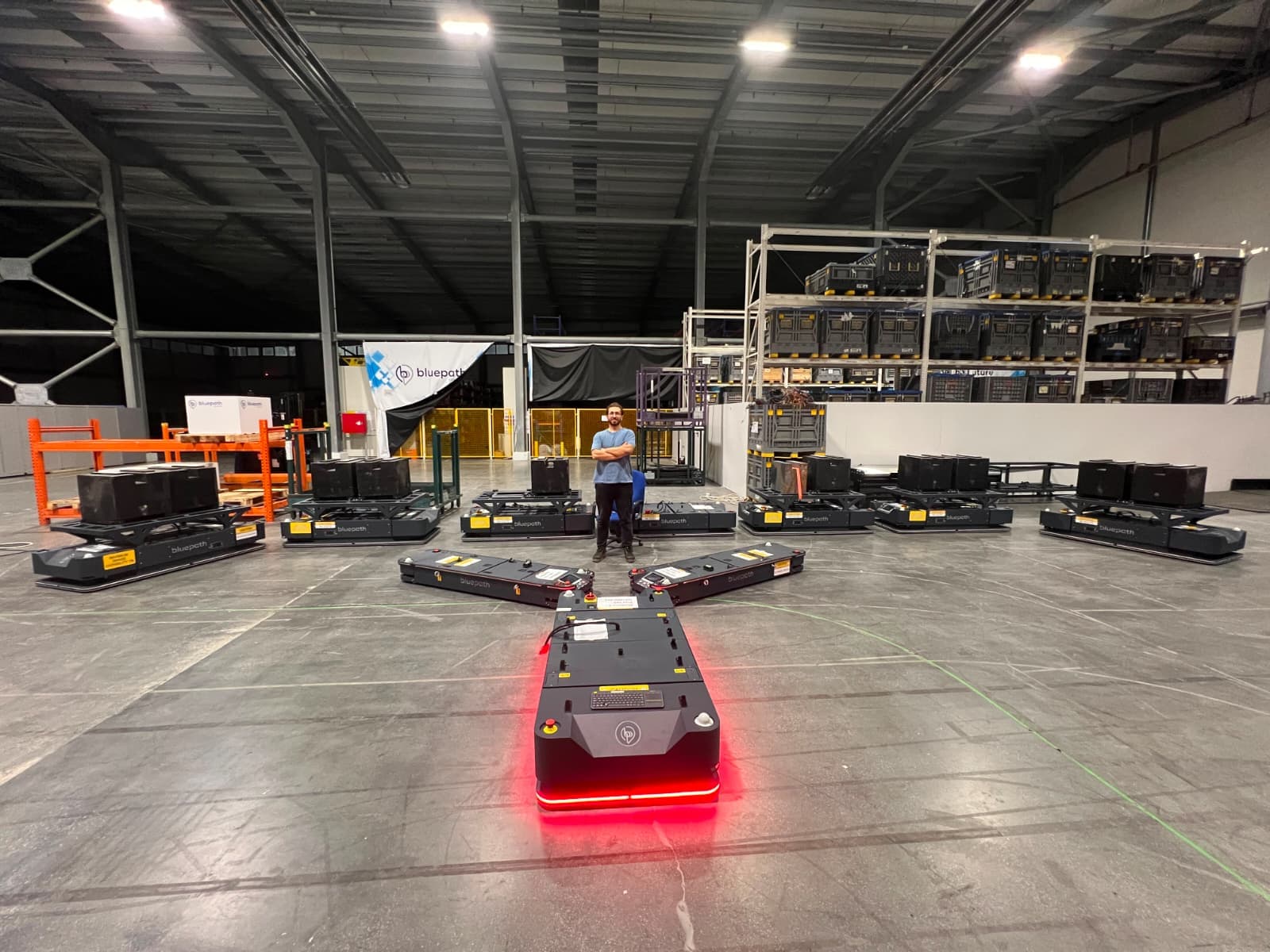 Tolgahan Gergin, depo test alanında yıldız düzeninde dizilmiş Bluepath otonom mobil robot filosunun ortasında